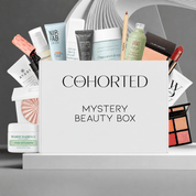 Cohorted Mystery Beauty Box set with a white box in the center and multiple beauty products fanned out behind it, including Nip+Fab, Dr. Brandit, Pixi, Laura Mercier, Mario Badescu, and Estée Lauder, on a gray studio background.