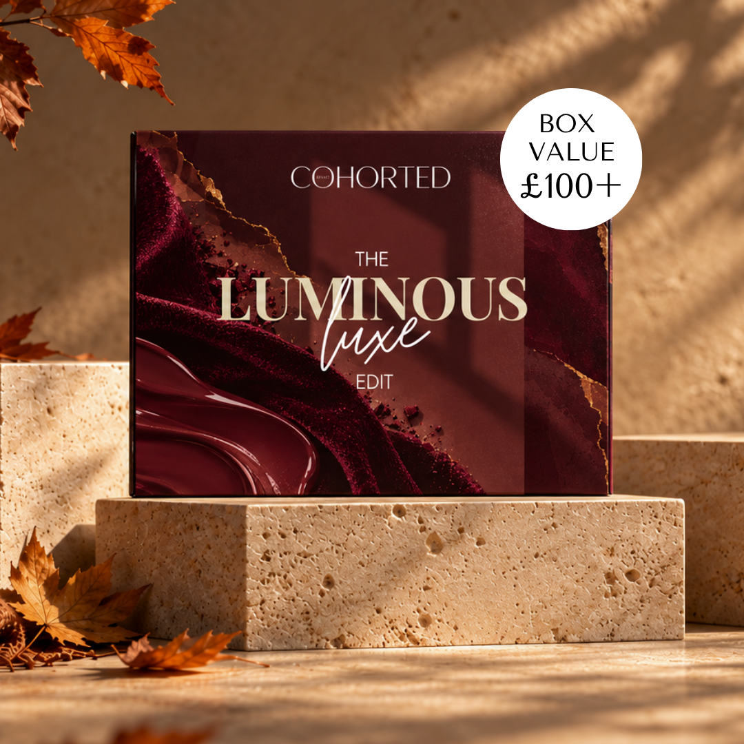 A boxed product labeled COHORTED The Luminous Luxe Edit stands on a stone pedestal in a warm, sunlit setting with autumn leaves around it. A white circular badge in the top right says BOX VALUE £100+. Luxury subscription beauty box, UK, gift for her